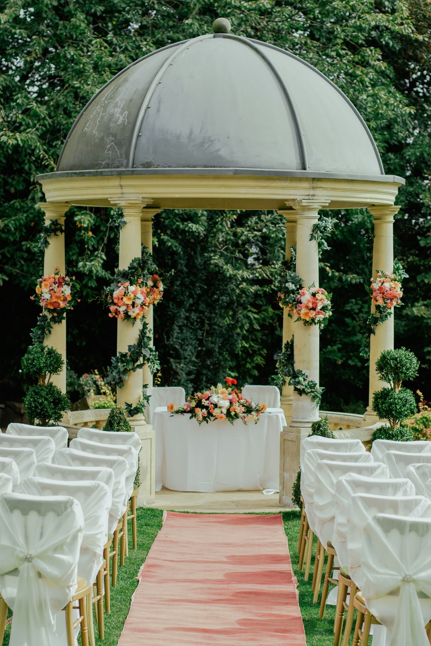 A garden wedding aisle with white chairs leading to a flower-covered gazebo